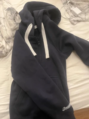 Mörkblå zip-hoodie från Russemerch  - Mörkblå hoodie från Russemerch med dragkedja framtill och stora vita snören i huvan. Mjuk insida i fleece och känguruficka framtill. Dock 2 Blekningsfläckar som nog ej går bort! Men syns knappt eftersom det är i luvan.  Klassisk och enkel design som passar perfekt till jeans eller mjukisbyxor.