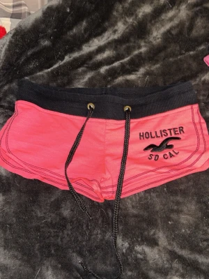 Rosa shorts från Hollister med snörning - Snygga rosa shorts från Hollister med svart midjeresår och snörning framtill. Hollister-logga och text broderat på ena benet. Perfekta för sommaren och chill dagar. Mjukt material och sportig look.