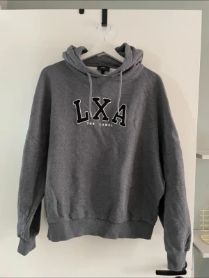 Grå hoodie från LXA The Label - Mysig grå hoodie från LXA The Label med stor huva och dragsnören. Framtill finns ett broderat LXA-tryck i svart och vitt. Hoodien har ribbade muddar vid ärmslut och nederkant, samt en avslappnad passform. Perfekt för en chill och casual stil.