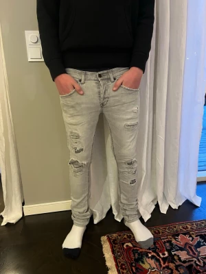 Grå dondup - Säljer ett par grå skinny jeans med slitningar och hål på framsidan. Jeansen har klassisk femficksdesign och är i stretchigt denimtyg för skön passform. Perfekta för dig som gillar en edgy och avslappnad stil. 