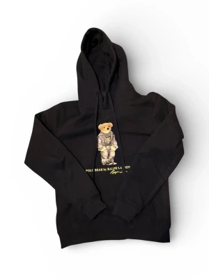 Blå hoodie Polo Bear Ralph Lauren - Blå hoodie från Ralph Lauren med Polo Bear-tryck på bröstet. Klassisk huva med dragsko och ribbade muddar. Mjuk bomullskänsla och avslappnad passform. Perfekt för dig som gillar streetstyle och ikoniska motiv.