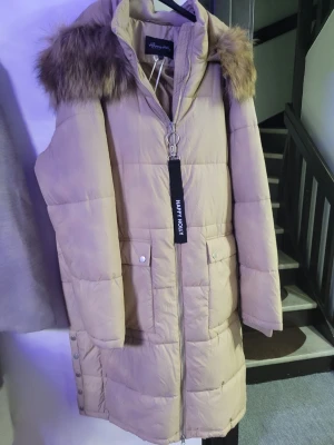 Beige lång dunjacka Happy Holly - Lång beige dunjacka från Happy Holly med stor huva och fuskpälsdetalj. Jackan har dragkedja framtill, två fickor med tryckknappar och dekorativt band vid dragkedjan. Perfekt för kalla vinterdagar och riktigt mysig att bära.