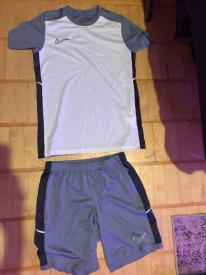 Nike grått träningsset t-shirt & shorts - Säljer ett sportigt träningsset från Nike med grå t-shirt och matchande shorts. T-shirten har vita och svarta detaljer samt Nike-logga på bröstet. Shortsen är grå med svarta och vita inslag och har elastisk midja. Perfekt för fotboll eller gym.