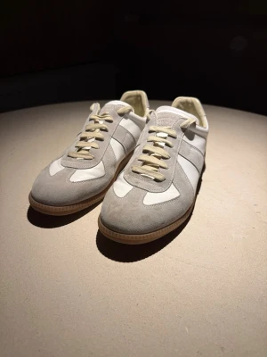 Maison Margiela vit och grå - Snygga sneakers i vitt och ljusgrått med ovandel i både mocka och skinn. Klassisk låg modell med beige snören och gummisula. Perfekta för en clean och stilren look.