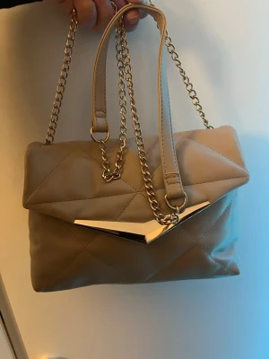 Väska - Snygg beige handväska i quiltat mönster med guldfärgade kedjehandtag och en cool metallspets framtill. Väskan har en mjuk, rektangulär form och stängs med ett lock. Perfekt accessoar för att lyfta din outfit. Köptes för 200kr