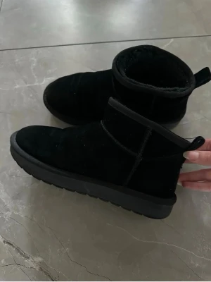 Svarta UGG liknande skor i mocka - Säljer ett par svarta UGG liknande boots i mjuk mocka med låg ankelhöjd och tjock sula. Insidan är fodrad med fluffigt material för extra komfort. Klassisk design med dragflik bak. Perfekta för att hålla fötterna varma under kalla dagar.