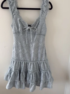 Ljusblå spetsklänning med volang - Supersöt ljusblå kortklänning i spets med volangkant och knyt framtill. Klänningen har breda axelband och v-ringning, samt ett romantiskt mönster i tyget. Perfekt för sommarens alla tillfällen!