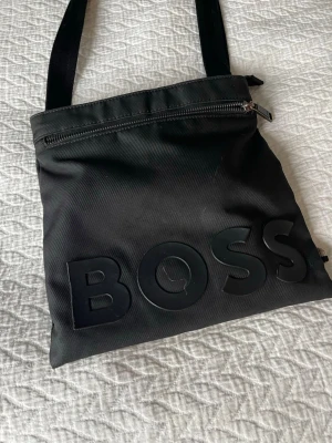 Svart axelväska från BOSS - Snygg svart axelväska från BOSS med stort logotyptryck framtill. Väskan har justerbar axelrem och en dragkedja upptill. Tillverkad i slitstarkt tygmaterial och har ett rymligt huvudfack. Perfekt för att bära det viktigaste med stil.