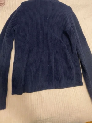 Mörkblå stickad tröja med dragkedja - Mörkblå stickad tröja med halv dragkedja framtill och ståkrage. Tröjan har lång ärm och är ribbstickad för en snygg och enkel look. Perfekt för lager-på-lager och passar till det mesta.