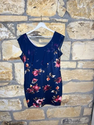 Mörkblå blommig klänning med volang - Mörkblå klänning med stora rosa och röda blommor. Klänningen har en bred volang över axlarna och en djupare rundringning både fram och bak. Ärmlös modell i mjukt, lätt material