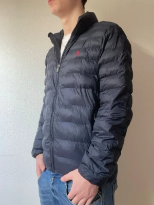 Mörkblå dunjacka från Ralph Lauren - Snygg Mörkblå dunjacka från Ralph Lauren med diskret röd logga på bröstet. Jackan har dragkedja framtill, ståkrage och två sidofickor. Lätt och smidig modell som håller värmen bra under kalla dagar. Passar perfekt till jeans och t-shirt. Modellen är 178cm!