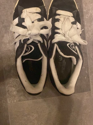 Adidas Campus svartvita sneakers - Säljer ett par klassiska Adidas Campus sneakers i svart mocka med vita detaljer och de ikoniska tre ränderna på sidan. Skorna har vita snören och en låg siluett som ger en clean och tidlös vibe. CAMPUS-loggan syns på sidan. Perfekta för en streetwear-look.