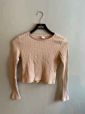 Beige ribbad croppad topp - Trendig croppad topp i beige med ribbad struktur och långa ärmar. Perfekt till höga jeans eller kjol för en clean och enkel look. Mjuk och stretchig, passar dig som gillar stilrena basplagg. Pris är absolut diskuterbart