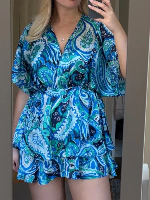 Blå paisleymönstrad kortklänning - Kort klänning med blått paisleymönster i olika nyanser av blått, grönt och vitt. Klänningen har v-ringning, volangdetaljer vid kjolen och lös passform med resår i midjan. Tillverkad i ett lätt och luftigt material, perfekt för sommaren🫶🏼