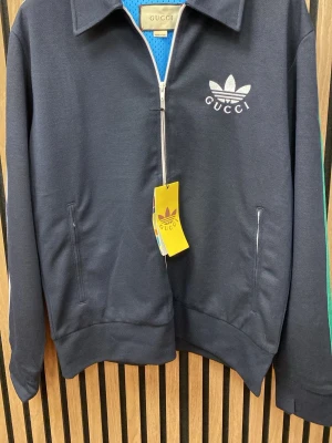 Marlinblå vindjacka  - Snygg svart vindjacka med vit dragkedja, klassisk krage och logga på bröstet. Jackan har långa ärmar med gröna och röda ränder på sidan samt fickor med dragkedja. Perfekt för dig som gillar streetwear och exklusiva samarbeten.