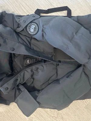 Mörkgrå jacka från Canada Goose - Säljer en svart dunjacka från Canada Goose med klassisk patch på ärmen. Jackan har huva, dragkedja och tryckknappar framtill. Tjockt vadderad för extra värme och perfekt för kalla vinterdagar. Snygg och tidlös design som funkar till det mesta. ( WYNDHAM PARKA) kvitto finns med 