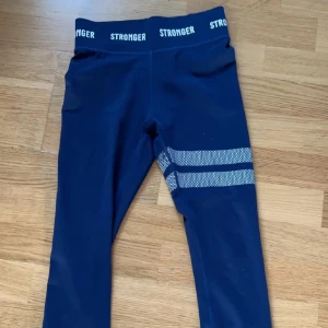 Blå leggings från Stronger - Mörkblå leggings från Stronger med bred resår i midjan där märkesnamnet upprepas i vitt. Snygg detalj med två vita, prickiga ränder på ena benet. Stretchigt material som sitter skönt och följsamt.