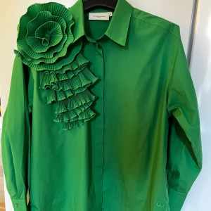 Grön blus med volangdetaljer från Copenhagen Muse - Säljer en grön säblus från Copenhagen Muse med unika volangdetaljer (blomma) i plisserat tyg över ena axeln. Blusen har klassisk krage och långa ärmar. Perfekt för dig som vill sticka ut med en färgstark och cool look. Storleken är S m passar mig som är 38/40. Använd 2 ggr. Nypris 1200 kr