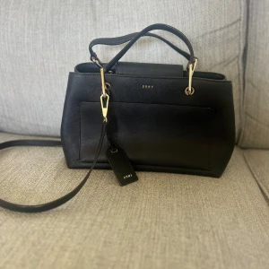 Svart handväska från DKNY - Snygg svart handväska från DKNY med guldfärgade metalldetaljer och avtagbar axelrem. Väskan har flera fack, dragkedja på insidan och en stilren design. Perfekt för dig som vill ha en klassisk och tidlös accessoar.