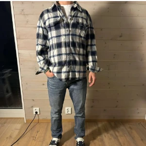 Flanellskjorta  - Säljer en blåvit rutig overshirt med långa ärmar och klassisk krage. Skjortan har knappar framtill och en bröstficka. Perfekt att slänga över en t-shirt för en avslappnad look.