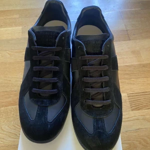 Mason Margiela - Säljer ett par svarta sneakers från Maison Margiela med snygga mockadetaljer och klassisk design. Skorna har svarta snören, rund tå och platt sula. Perfekta för dig som gillar stilrena och tidlösa sneakers.