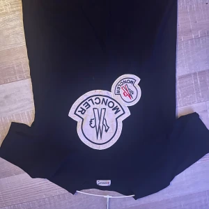 Svart t-shirt från Moncler med stora logopatchar - Svart t-shirt från Moncler med två stora vita logopatchar på bröstet, en med svart och en med röd detalj. Klassisk rund halsringning och korta ärmar. Perfekt för dig som gillar streetwear och tydliga märkesdetaljer. Är stolek M men är mer som S
