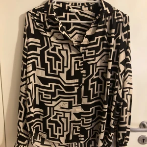Svartvit mönstrad skjorta från Richard Allan x H&M - Säljer en svartvit skjorta med grafiskt mönster från Richard Allan x H&M. Skjortan har klassisk krage, knäppning framtill och långa ärmar. Perfekt för dig som gillar unika prints och vill sticka ut.
