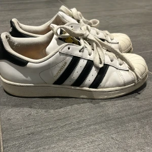 Adidas Superstar sneakers i vitt och svart - Klassiska Adidas Superstar sneakers i vitt skinn med svarta ränder och gulddetaljer på plösen. Några defekter på insidan av skon som syns på bild fyra. Annars bra skick. Skicka gärna frågor. Pris går att diskutera 