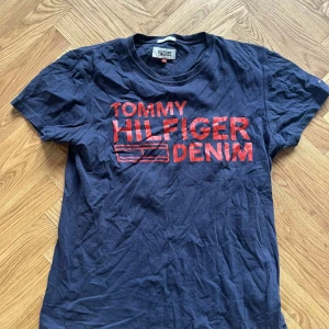 Mörkblå t-shirt från Tommy Hilfiger  - Säljer en mörkblå t-shirt från Tommy Hilfiger Denim med röd och vit logga tryckt på bröstet. Klassisk passform och rund halsringning. Perfekt för en avslappnad stil.