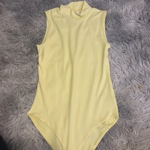 Ljusgul body ärmlös från H&M - Säljer en ljusgul ärmlös body från H&M. Toppen är ribbad och har en hög krage, perfekt för att styla med jeans eller kjol. Mjuk och stretchig i materialet för en bekväm passform.