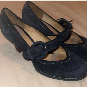 Navy Mary Janes - Säljer ett par mörkblå pumps i mocka med bred rem och stor rund spänne över foten. Skorna har rund tå och medelhög klack, samt dekorativa sömmar längs kanterna. Perfekta för dig som gillar klassisk och stilren design.💙