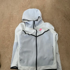 Vit hoodie från Nike med svarta detaljer - Nike hoodiejacka i vitt med svarta kontrastdetaljer och dragkedja. Jackan har huva och en rosa Nike-logga på bröstet. Sportig design med fickor framtill och lång ärm. Perfekt för dig som gillar streetwear och sportig stil.