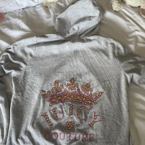 Grå hoodie från Juicy Couture med krona och strass - Grå zip-hoodie från Juicy Couture med huva och fickor framtill. Baksidan har en stor krona dekorerad med rosa och guldiga strasstenar samt texten 'Juicy Couture'. Väldigt fin och Y2K som jag älskat såååå mycket. Tyvärr har jag mer eller mindre tröttnat på den… nypris är 2999 men jag köpte på outlet för 2000. Köparen står för frakten om inget annat sägs❤️