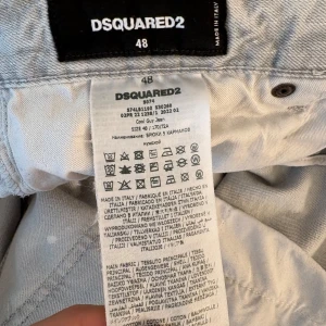 DSQUARED2 jeans  - Säljer ett par äkta  ljusgrå jeans från DSQUARED2 med coola slitningar och färgstänk. Jeansen har klassisk femficksdesign, knappgylf och en avslappnad look. Perfekta för dig som gillar streetstyle och vill sticka ut lite extra.