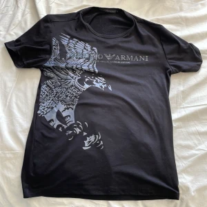 Svart t-shirt med örntryck från Emporio Armani - Snygg svart t-shirt från Emporio Armani med stort grått örntryck och logga på bröstet. Klassisk passform och rund halsringning. Perfekt för dig som vill ha en stilren men ändå iögonfallande look. Säljer eftersom den inte passar mig längre, inga defekter. Priset är ej fast och kan gärna skicka fler bilder !! 