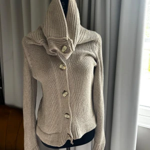 Tröja med knappar - Säljer en beige stickad tröja med unik stor krage och fyra stora knappar framtill. Koftan har lång ärm och är ribbstickad i mjukt material. Perfekt för vardag och en cool detalj till din outfit.