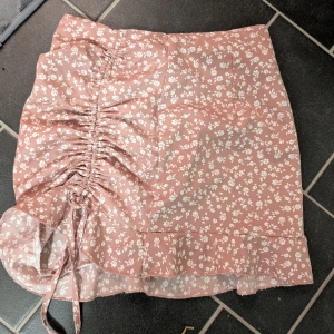 Rosa blommig kjol med volang och snörning - Supersöt rosa kjol med vitt blommönster. Kjolen har volang nertill och snygg snörning på sidan som ger en rynkad effekt. Perfekt för dig som gillar romantisk och trendig stil. Använd fåtal gånger