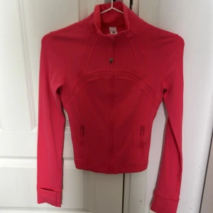 Rosa träningsjacka från Lululemon - Säljer en snygg rosa träningsjacka från Lululemon i storlek 2 (XS). Jackan har dragkedja framtill, hög krage och två fickor med dragkedja. Perfekt för träning eller som ett sportigt lager.