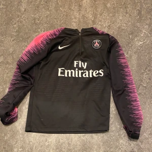 Svart långärmad Paris Saint-Germain fotbollströja från Nike - Säljer en svart långärmad PSG-tröja från Nike med rosa detaljer längs ärmarna och axlarna. Tröjan har halv dragkedja framtill, Paris Saint-Germain-logga på bröstet och Fly Emirates-tryck. Tillverkad i Dri-FIT-material. Träningströja från 2018-19 säsongen.