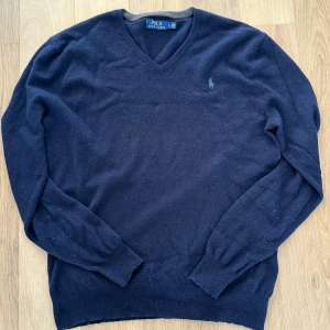 Mörkblå v-ringad tröja från Polo Ralph Lauren storlek Large - Mörkblå tröja från Ralph Lauren i storlek Large. Använd endast ett fåtal gånger och är i mycket fint skick – ser nästan ny ut. Klassisk modell med V-ringning och broderad logotyp på bröstet. Normal passform.