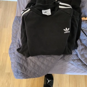 Svart sweatshirt från Adidas med vita ränder beg skick - Svart sweatshirt från Adidas med klassiska vita ränder på axlarna och Adidas logga på bröstet. Tröjan har rund halsringning och långa ärmar. Perfekt för en sportig och avslappnad stil. Beg skick Stl L