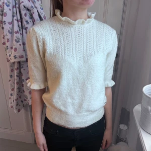 Vit stickad tröja  - Säljer en söt vit stickad tröja med korta ärmar och volangdetalj vid halsen och ärmsluten. Tröjan har ett diskret hålmönster upptill och ribbade muddar. Perfekt för dig som gillar en romantisk och stilren look.