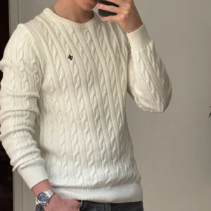 Vit kabelstickad tröja från Morris - Säljer en klassisk vit kabelstickad tröja från Morris. Tröjan har rund halsringning, ribbade muddar och ett diskret broderat emblem på bröstet. Perfekt för en stilren och tidlös look.