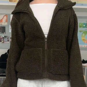 Mörkgrön teddyjacka med dragkedja - Mysig mörkgrön teddyjacka med dragkedja framtill och stora fickor. Jackan har en bred krage och är långärmad. Perfekt för kyligare dagar och ger en avslappnad look.