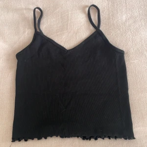Svart ribbad linnetopp från H&M - Säljer en svart ribbad linnetopp från H&M med smala axelband och v-ringning. Toppen har en croppad passform och vågig kant nertill. Perfekt att styla med jeans eller kjol. Jag kommer tvätta produkten innan jag skickar den. 