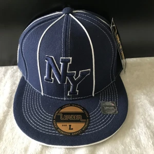 Mörkblå keps med vita detaljer från Leader - Snygg mörkblå keps med vita kontrastsömmar och broderad NY-logga framtill. Kepsen har platt skärm och justerbar passform. Perfekt för dig som gillar sportig stil. Finns S ,L stlk kvar  109kr /st 