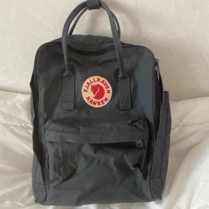 Grå ryggsäck från Fjällräven Kånken - Klassisk grå ryggsäck från Fjällräven Kånken med det ikoniska runda röda logotypmärket framtill. Väskan har ett stort huvudfack, en mindre ficka med dragkedja på framsidan och två handtag upptill.