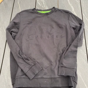 Svart sweatshirt från Hugo Boss - Säljer en svart sweatshirt från Hugo Boss med diskret broderad logga framtill. Tröjan har rund halsringning, ribbade muddar och en strukturerad baksida. Slim fit-modell med snygga detaljer och klassisk stil.