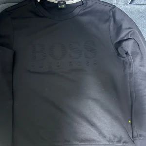 Svart sweatshirt från BOSS - Säljer en svart sweatshirt från BOSS med diskret logotyp i samma färg på bröstet. Tröjan har rund halsringning och långa ärmar. Passformen är slim fit och materialet känns mjukt och bekvämt.