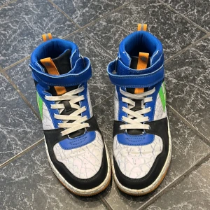 Färgglada höga sneakers med blå, grön och orange detalj - Säljer ett par höga sneakers med vit bas och svarta, blå och gröna partier samt orange detaljer. Skorna har snörning, kardborreband och mönstrad ovandel. Perfekta för dig som gillar färg och unika skor! Kan förhandlas/fråga gärna frågor!
